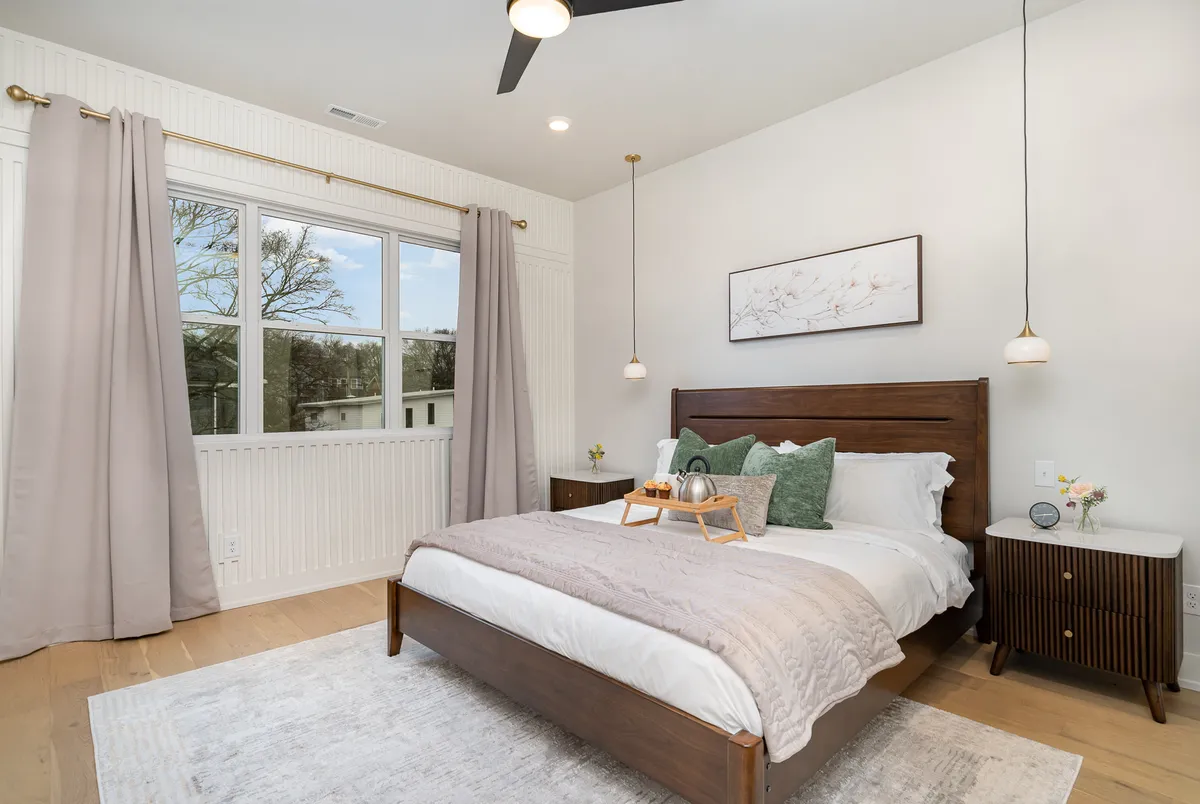 A serene bedroom with a neatly made bed adorned with decorative pillows, flanked by nightstands and lamps, and natural light streaming in through a window with curtains. The space feels inviting and restful, ideal for unwinding after a busy day.