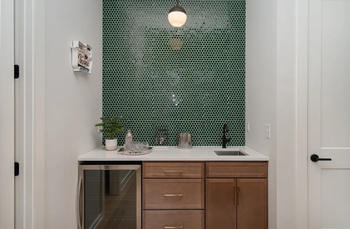 A stylish wet bar area boasting a green hexagonal backsplash, wood cabinetry, modern faucet, and glassware on the counter. The mood is welcoming and ideal for cocktail hour or casual celebrations with friends.